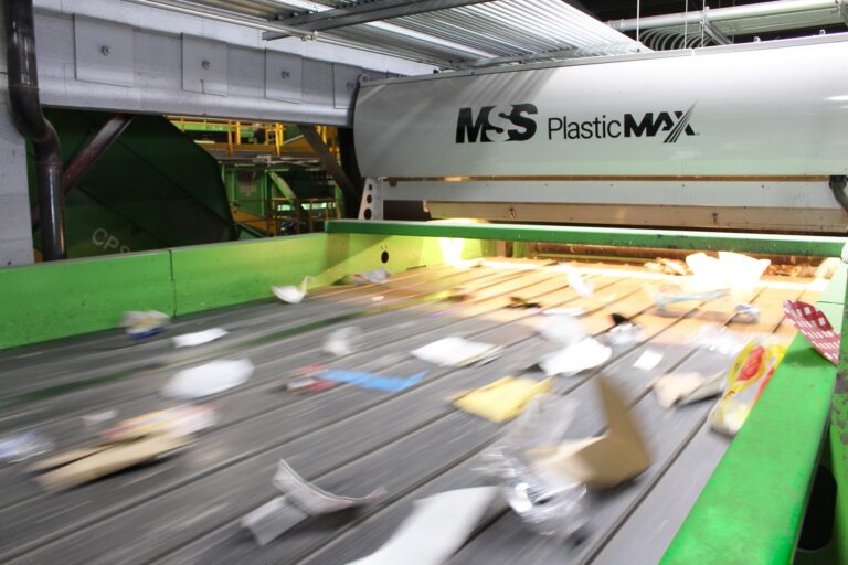 A sorting conveyor belt in a recycling facility, with various items of plastic waste moving along it. A machine labeled "MSS PlasticMAX" is in the background, indicating a focus on sorting plastic materials. The setting is industrial with green accents.