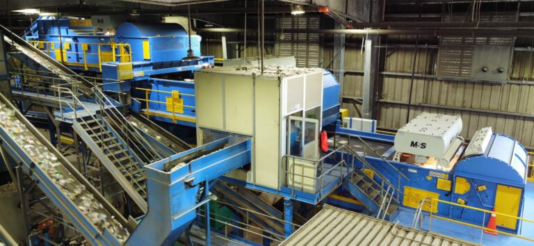 A recycling facility with conveyor belts and machinery sorting through materials. The setup includes multiple blue conveyor systems, a control booth, and various equipment used for processing and separating recyclables within a large industrial space.