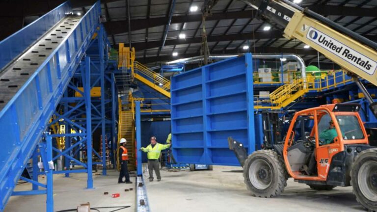 Workers in safety gear guide a forklift that is lifting a large blue metal structure inside an industrial MRF facility. There are blue conveyor belts and yellow railings in the background. The forklift has a "United Rentals" sign.