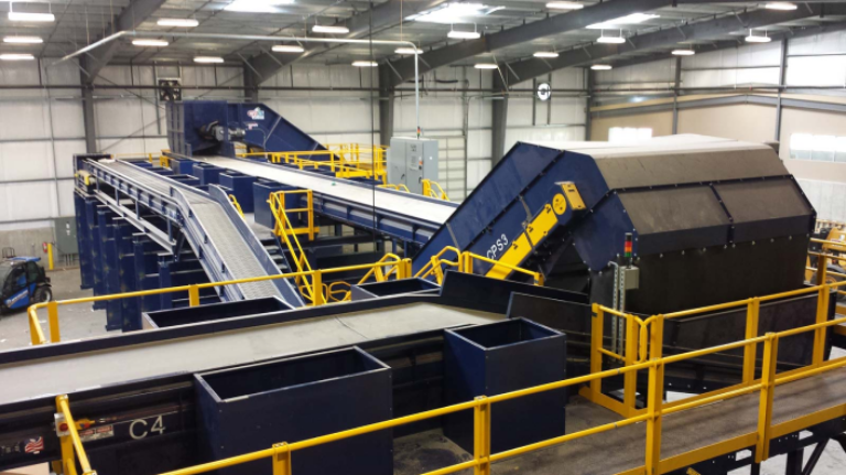 A modern recycling facility interior with large conveyor belts, sorting machinery, and elevated walkways. The McMRF equipment is primarily blue with yellow railings and structures. The space is brightly lit with industrial lighting overhead.