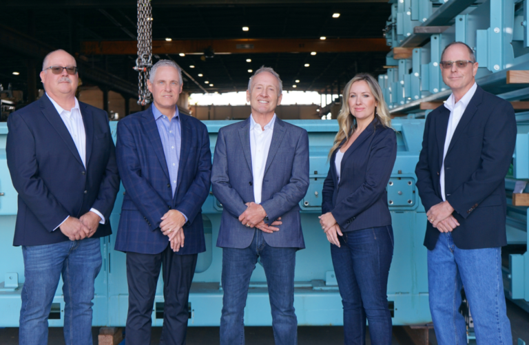 Five individuals stand in a row, posing for a group photo in a warehouse. They are dressed in business attire, and industrial equipment is visible in the background. Four men in dark blazers and jeans, and one woman in a dark blazer and jeans, are smiling at the camera.