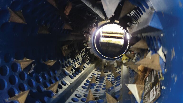 An interior view of a blue cylindrical machine with various angled metal blades and perforated sections. Light beams through an opening at the far end, illuminating parts of the circular structure and its mechanical components.