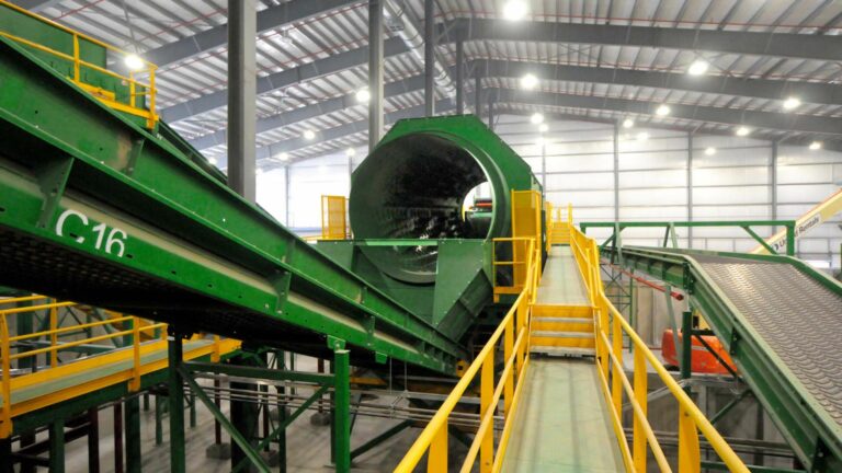 A large industrial facility with green machinery, including conveyor belts and a cylindrical rotating drum. The equipment is elevated with yellow safety rails, and the interior of the building has a high ceiling with supportive steel beams.