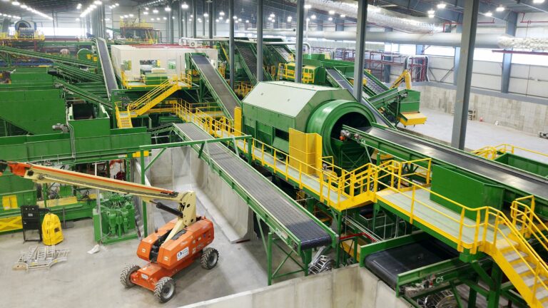 A large indoor waste management facility with various green machinery and conveyor belts processing materials. A cherry picker is positioned on the ground below, and bright overhead lights illuminate the space. The facility has high ceilings and an industrial design.