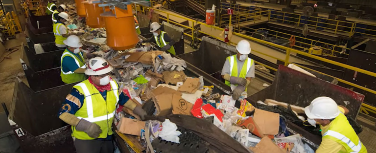 Workers in safety vests, helmets, and masks sort through a stream of mixed recyclables on a conveyor belt in a recycling facility. The background shows industrial equipment and other workers performing similar tasks.