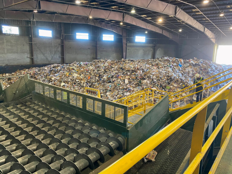 A large indoor facility with high metal ceilings, filled with heaps of mixed waste and recyclables. In the foreground, there is an Auger Screen sorting machinery with large metal rollers. A person in a safety vest stands near the machinery, overseeing the sorting process.