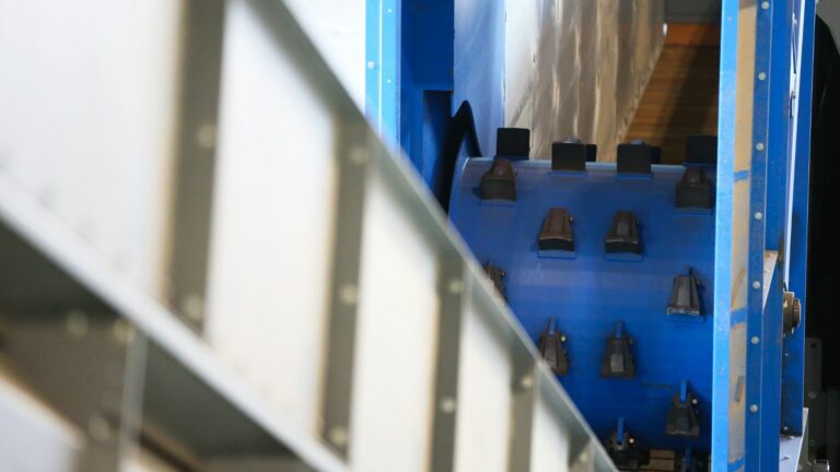 Close-up of a Drum Feeder machine with a conveyor belt on the left and multiple small components aligned on a blue background on the right. The image features a mix of metallic and textured surfaces, suggesting a manufacturing or assembly environment.