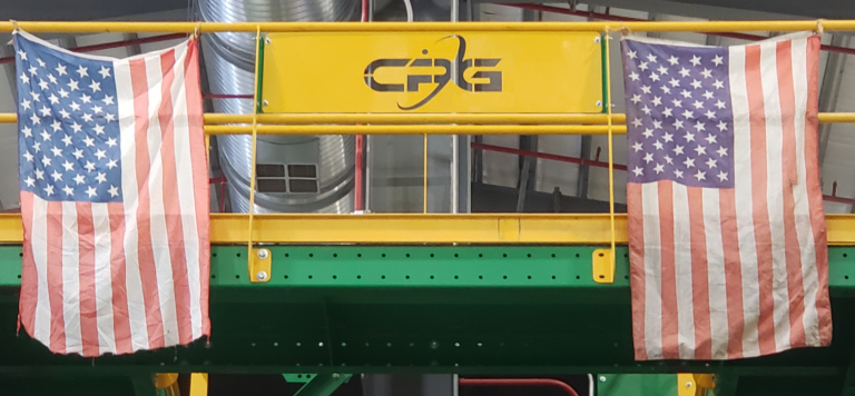 Two worn American flags hang from a yellow railing with the letters "CPG" on it. The background features industrial elements, including pipes and ducts, painted green and red. The setting appears to be inside a factory or large facility.