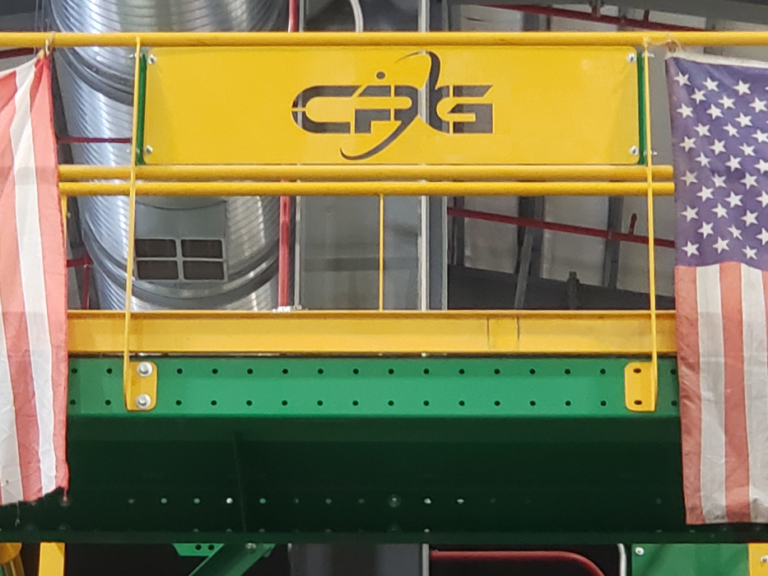 Two worn American flags hang from a yellow railing with the letters "CPG" on it. The background features industrial elements, including pipes and ducts, painted green and red. The setting appears to be inside a factory or large facility.