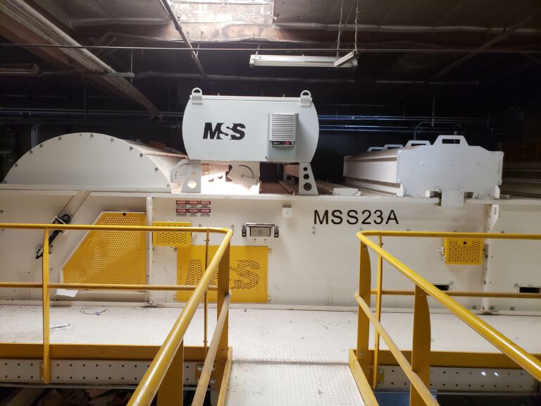 Sorting equipment labeled "MSS" and "MSS23A" in a MRF setting, featuring a large cylindrical component and guardrails painted yellow. The machinery is situated indoors with a ceiling and visible overhead lighting.