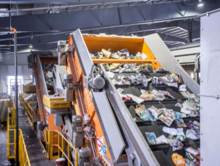 A large indoor recycling facility with conveyor belts moving various recyclable materials such as cardboard, paper, and plastic. The conveyors are angled upwards, carrying the materials towards machinery for sorting. The background shows industrial equipment and catwalks.