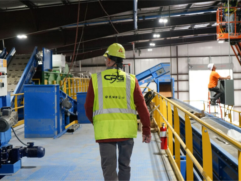 A person wearing a yellow safety vest and a hard hat is standing indoors in a warehouse-like setting. They are facing away, observing equipment and machinery. Another worker in an orange vest is elevated in a lift on the right side of the image.