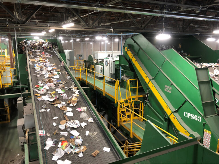 A large indoor recycling facility with several green conveyor belts sorting various materials such as paper, cardboard, and plastic. Overhead lights illuminate the machinery, and yellow railings provide safety along the platforms.