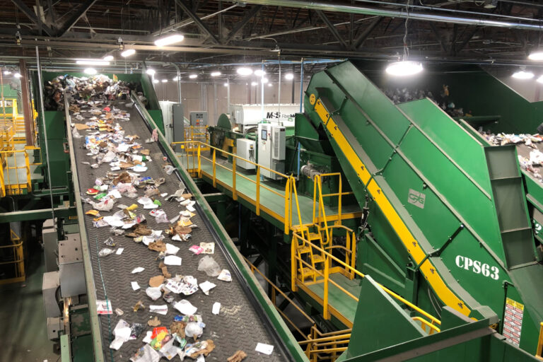 A large indoor recycling facility with several green conveyor belts sorting various materials such as paper, cardboard, and plastic. Overhead lights illuminate the machinery, and yellow railings provide safety along the platforms.