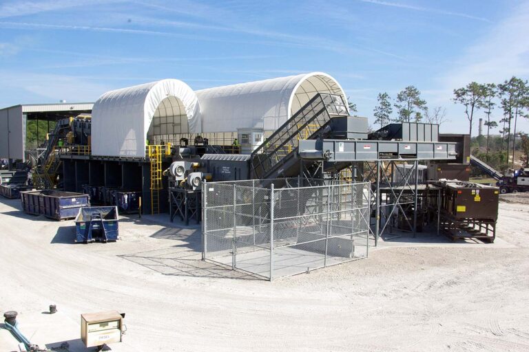 Industrial recycling facility with large white, arch-shaped structures and various equipment like conveyor belts. Metal fences surround some parts, and several large containers are scattered around. Trees and a clear blue sky are visible in the background.