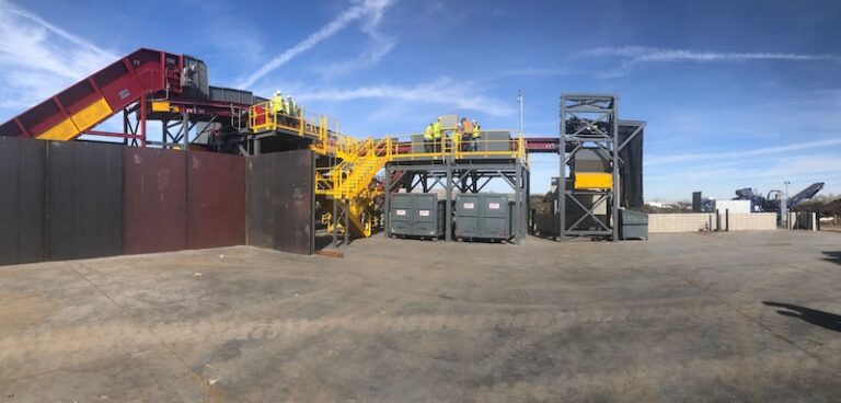 Industrial site with metal structures, a towering conveyor belt, and workers in safety gear. The background shows a clear blue sky with white clouds. The ground appears to be barren, and the overall environment is industrial and construction-oriented.