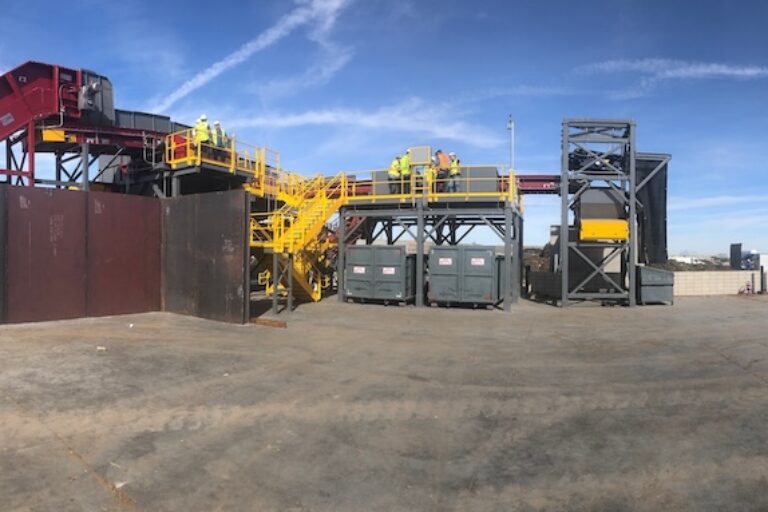 Industrial site with metal structures, a towering conveyor belt, and workers in safety gear. The background shows a clear blue sky with white clouds. The ground appears to be barren, and the overall environment is industrial and construction-oriented.