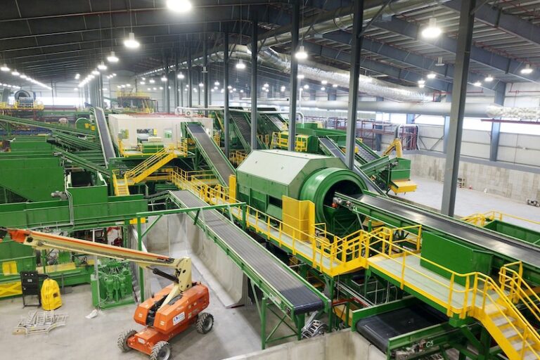 A large indoor MSW recycling facility with various green machinery and conveyor belts. A rotating drum and multiple sorting stations are visible. An orange boom lift is parked in the foreground. The setting is well-lit and spacious, with a high ceiling.