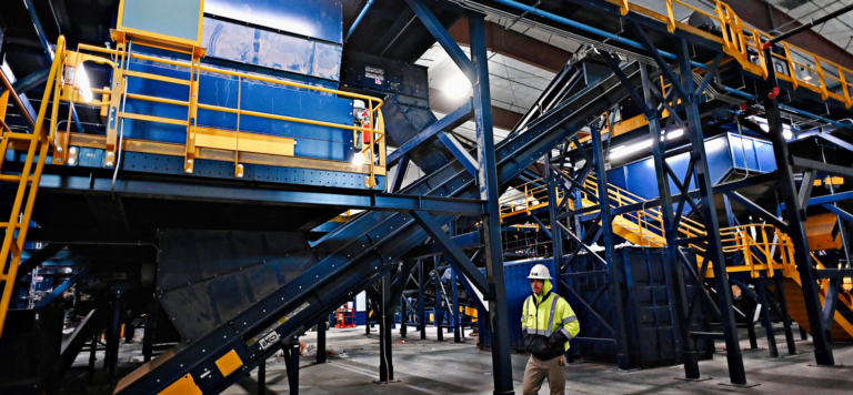 A worker in a yellow safety jacket and white hard hat stands in an industrial MRF filled with large blue and yellow machinery and conveyor belts. The area is well-lit with overhead lights and various metal frameworks and platforms are visible.