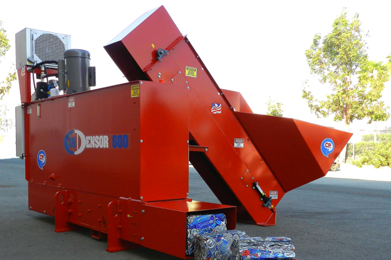 A large, red Can Densifier machine, labeled "CANDENSOR 600" is outdoors. It features a wide conveyor belt and appears to be processing and compacting bottles into bales. There are trees and a clear sky in the background.