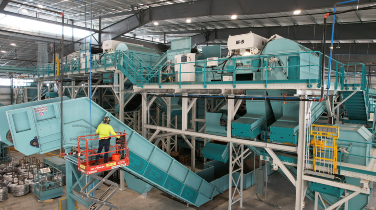 An industrial recycling facility with large machinery and conveyer belts. A worker in a yellow safety vest and helmet operates a small lift near a blue machine. Overhead lights hang from the ceiling, illuminating the large, complex equipment.