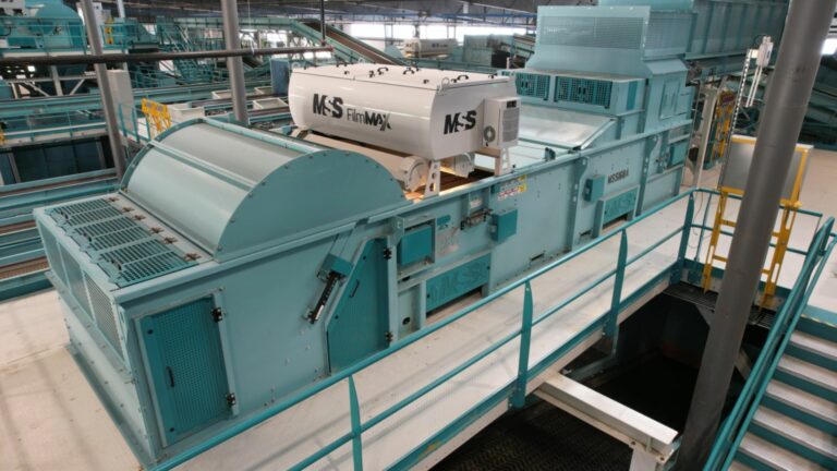 An Optical Sorting FilmMax machine in a large facility, featuring a long, light teal body with various components and piping. It is situated on a metal platform with railings and stairs for access, and has an "MSS" label on one section.