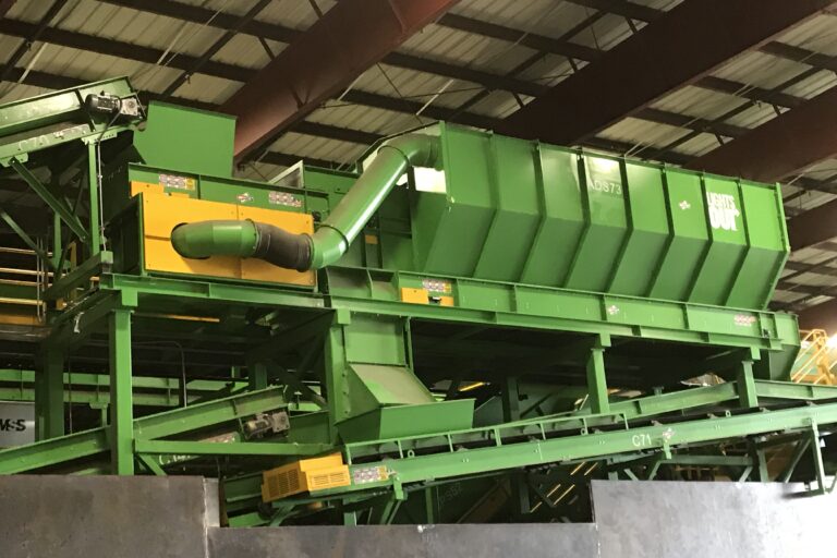 An industrial sorting machine with green metal parts and yellow highlights is inside a large warehouse. The machine has several conveyor belts and a chute, indicating its use in material processing or recycling operations.