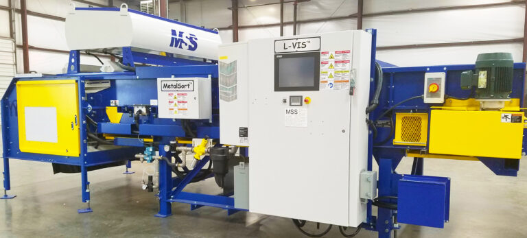 An L-VIS Optical Sorting machine featuring a large control panel with a screen at the center and multiple yellow and blue components on either side. The equipment is set in a spacious warehouse with high ceilings and exposed beams. Various cables and pipes are connected to the machine.