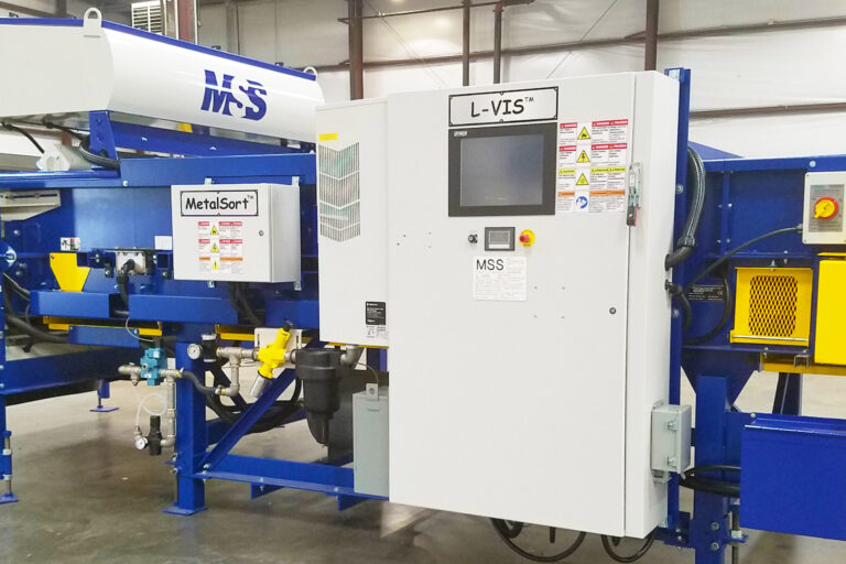 An L-VIS Optical Sorting machine featuring a large control panel with a screen at the center and multiple yellow and blue components on either side. The equipment is set in a spacious warehouse with high ceilings and exposed beams. Various cables and pipes are connected to the machine.