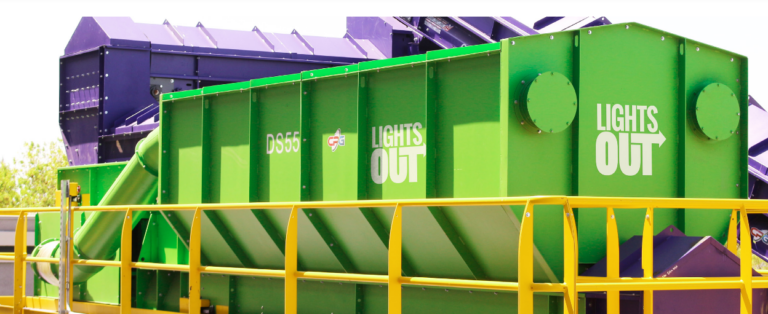 A bright green industrial machine with "Lights Out" written on the sides, surrounded by a yellow railing. The machine is set in an outdoor environment with another piece of machinery in the background.