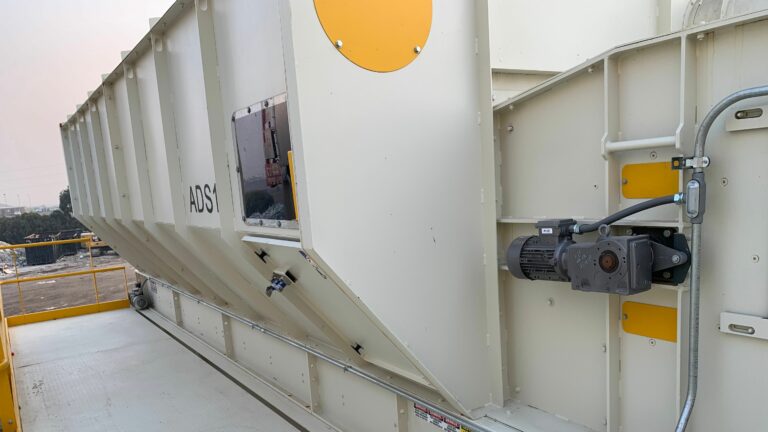 A large, industrial white metal structure with a motorized system and visible bolts, situated on a rooftop with some trees in the background. The unit is labeled "AOS1" and features vents and panels, likely used for ventilation or processing purposes.