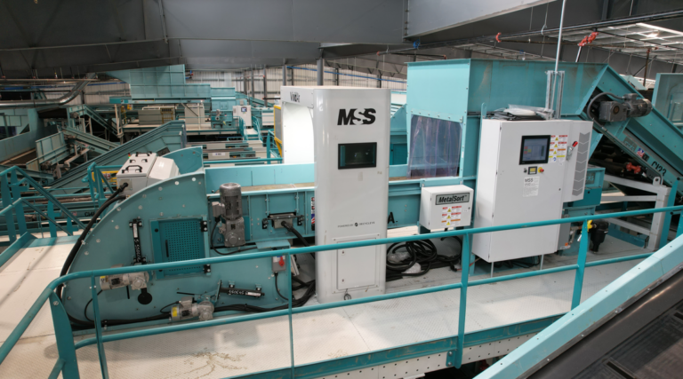 A MRF setting with a large, teal-colored Vivid AI sorting machine labeled "MSS". There are various control panels, conveyer belts, and wiring visible. The area is well-lit, and surrounding the machinery are metal railings on a white concrete floor.