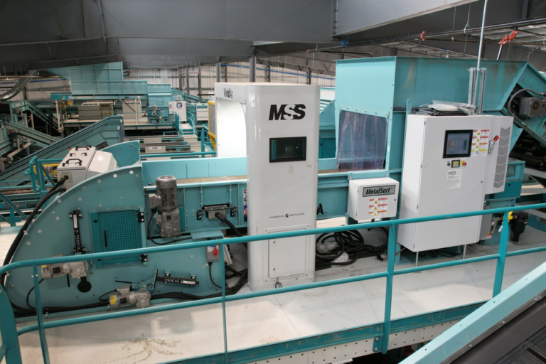 A MRF setting with a large, teal-colored Vivid AI sorting machine labeled "MSS". There are various control panels, conveyer belts, and wiring visible. The area is well-lit, and surrounding the machinery are metal railings on a white concrete floor.