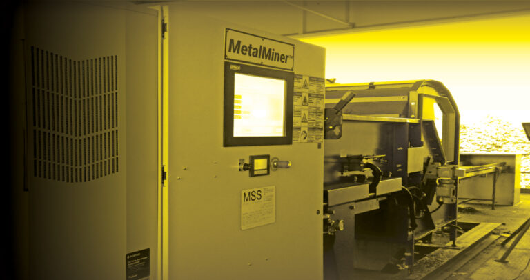 A yellow-tinted image featuring optical sorting machinery. In the foreground, there is a control panel labeled "MetaMiner" with a screen and buttons. Behind it, partially visible, is additional equipment, possibly part of a metal processing or manufacturing setup.