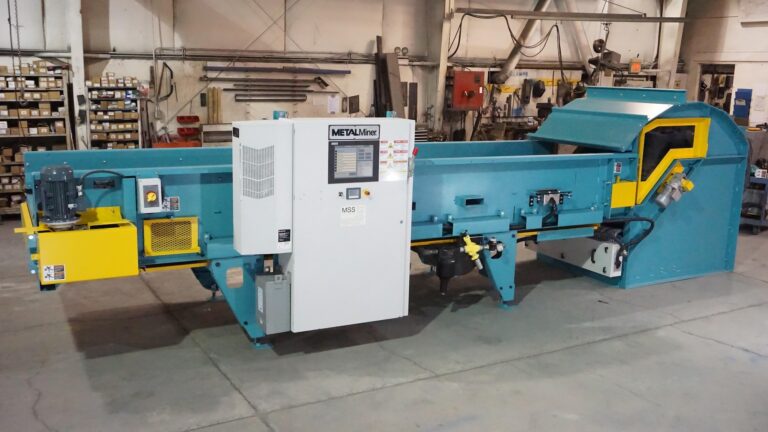 A large industrial sorting machine with a control panel, positioned in a spacious workshop. The machine features a teal body with yellow accents and various components for processing materials. Shelves and other equipment are visible in the background.