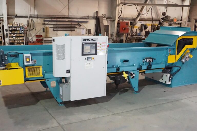 A large industrial sorting machine with a control panel, positioned in a spacious workshop. The machine features a teal body with yellow accents and various components for processing materials. Shelves and other equipment are visible in the background.