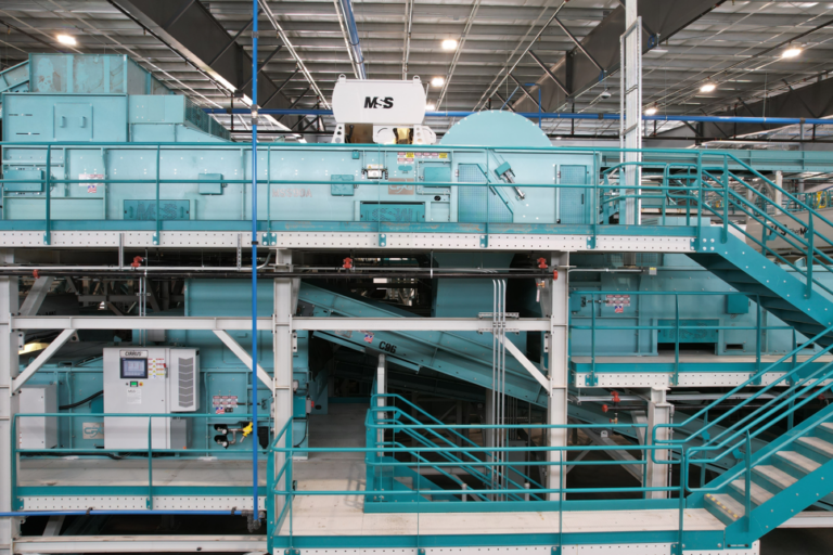 A large MRF facility features complex teal machinery with multiple optical sorters, staircases, and railings. Various levels and platforms are interconnected. Overhead signs label sections "MSS." The ceiling is lined with lights and visible structural elements.