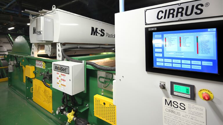 An industrial machine in operation inside a factory. A digital control panel labeled "CIRRUS" with various graphs and readings is prominently displayed. The PlasticMax machinery is green with labels such as "MSS" visible. The environment is clean and well-lit.