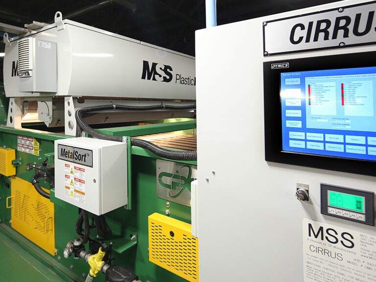 An industrial machine in operation inside a factory. A digital control panel labeled "CIRRUS" with various graphs and readings is prominently displayed. The PlasticMax machinery is green with labels such as "MSS" visible. The environment is clean and well-lit.