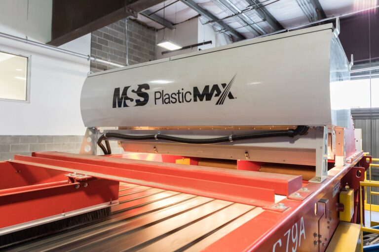 Close-up of industrial optical sorter labeled "MSS Plastic MAX" in a factory setting. The machine is mounted on a red frame, with a corrugated metal conveyor belt below it. The background features a gray brick wall, windows, and industrial lighting.