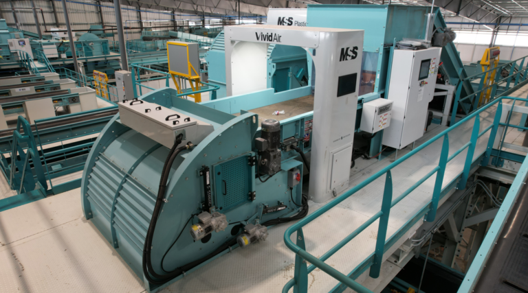 A modern MRF machinery setup inside a facility with teal-colored equipment. There's a large machine labeled "MSS" and "VividAI" with various controls and monitors. The environment is spacious and filled with similar equipment along an elevated walkway.