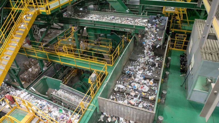 A vast waste processing facility with yellow railings and green machinery sorting through a large amount of mixed paper and plastic materials along conveyor belts. The scene is industrious, with multiple levels and bins filled with recyclables being processed.