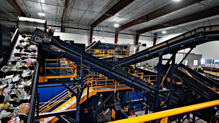 A Chicago recycling plant with multiple conveyor belts sorting various types of waste materials. The belts are intersecting and operating under a high ceiling with metal beams. Workers are visible in the background, managing and overseeing the process.