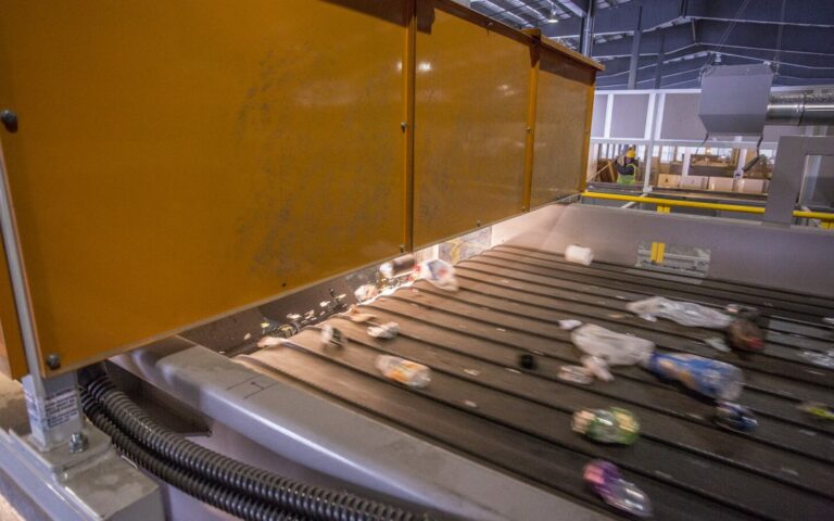 Conveyor belt with various items such as plastic bottles and containers moving underneath a large yellow machine in an industrial setting. The surroundings include metal structures and an industrial facility in the background.