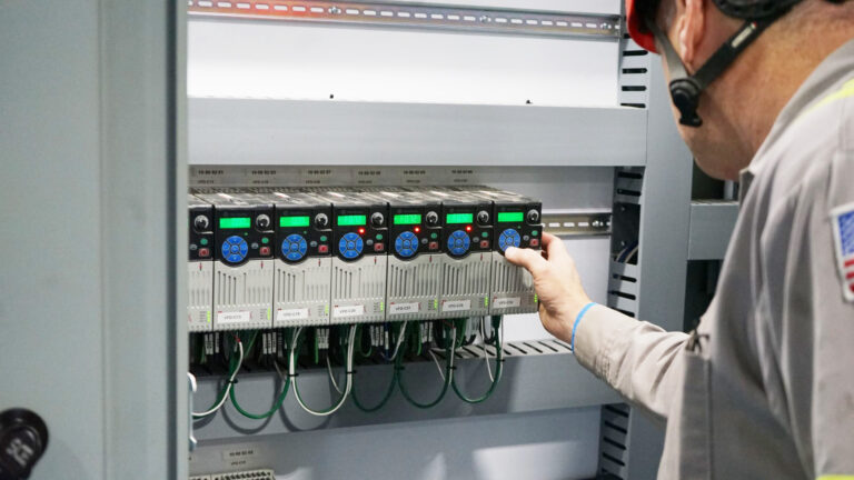 A technician in a gray uniform and a red helmet is adjusting controls inside an electrical panel with multiple electronic components and green and black wiring. The panel displays screens with illuminated indicators. The technician's face is not fully visible.