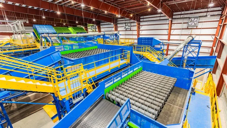 Recycling plant interior with blue conveyors, metal rollers and yellow safety railings beneath a high steel roof