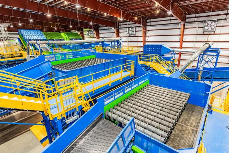 Recycling plant interior with blue conveyors, metal rollers and yellow safety railings beneath a high steel roof