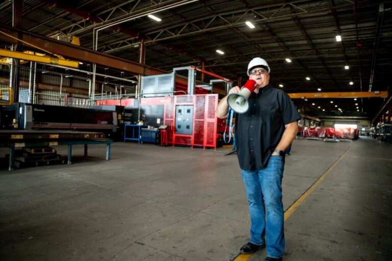 Man wearing white hard hat and safety glasses holding a red megaphone in a large industrial warehouse.