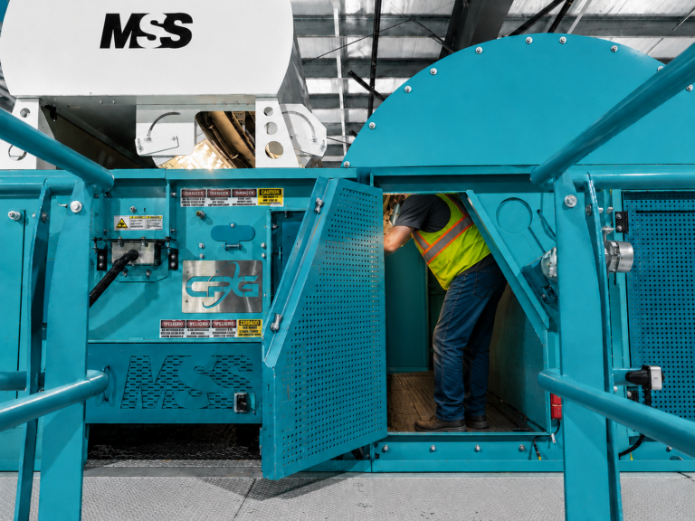 Worker in yellow safety vest and jeans leaning into open teal industrial machine access panel labeled MSS, factory setting.