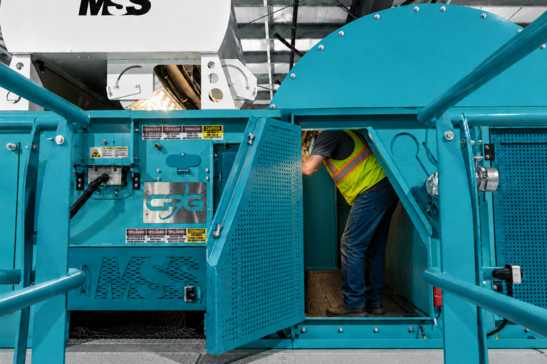 Worker in yellow safety vest and jeans leaning into open teal industrial machine access panel labeled MSS, factory setting.